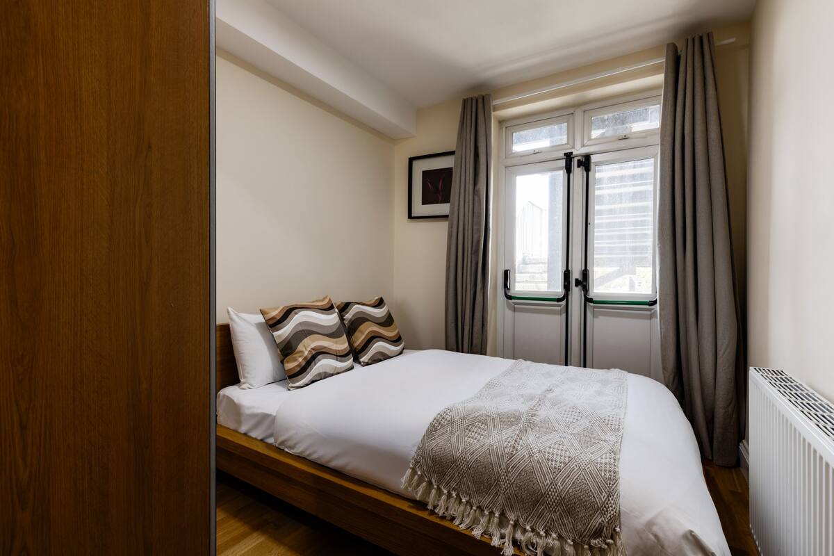 A bedroom with a bed, a window with curtains, a radiator, and a framed picture on the wall.