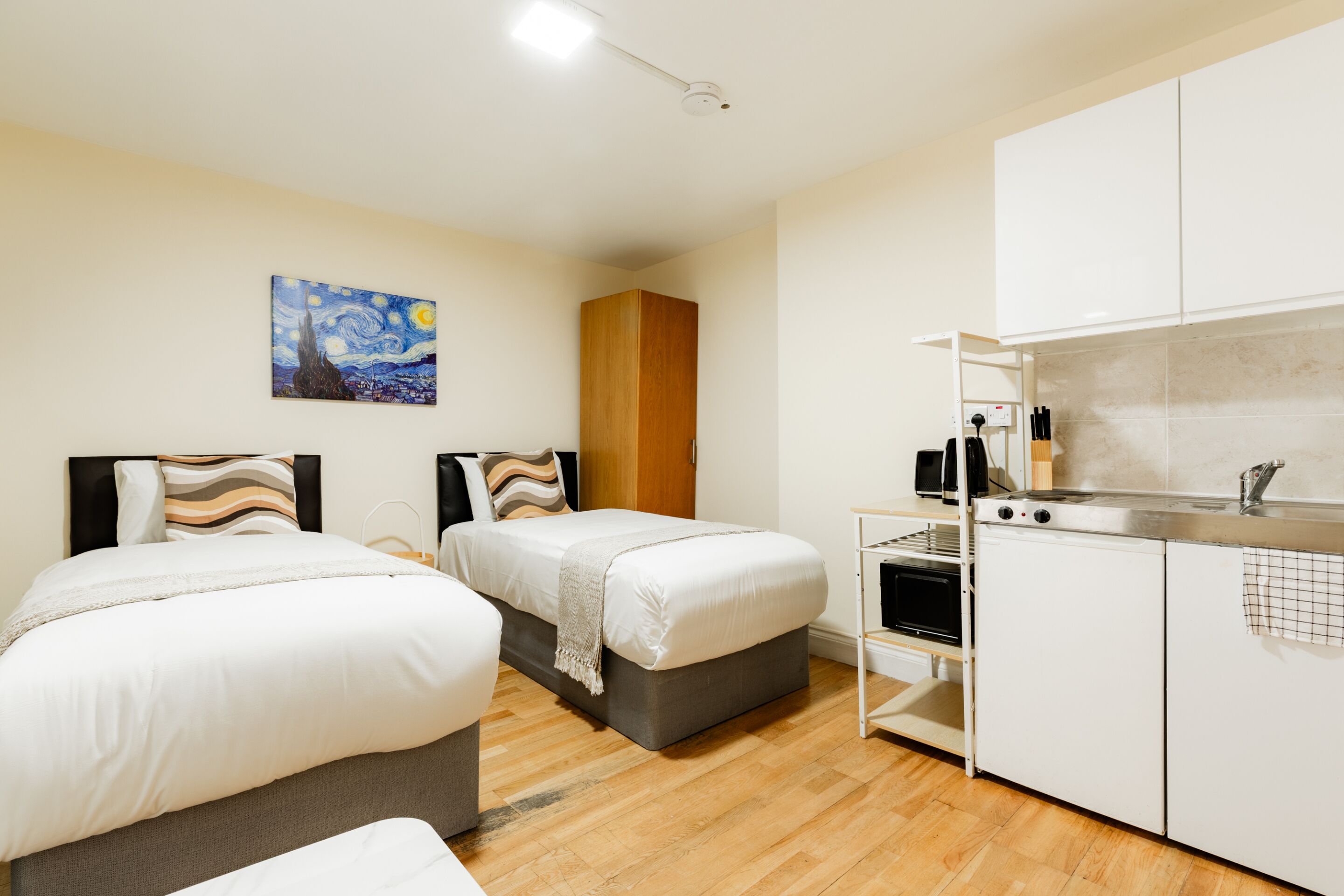 A hotel room with two beds, a kitchenette, and a painting on the wall.