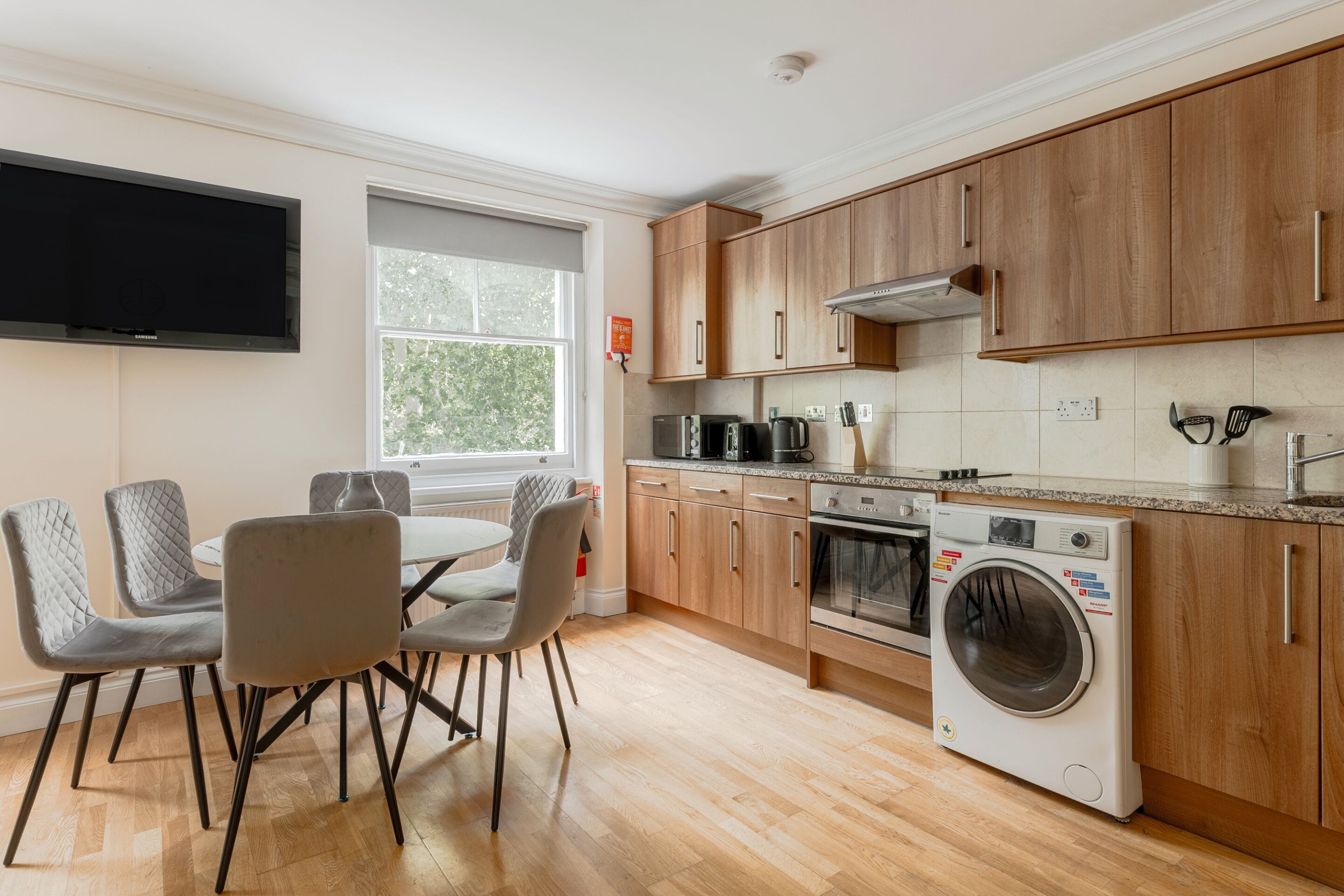 A modern kitchen with wooden cabinets, a built-in oven, a washing machine, and a dining area with grey chairs.