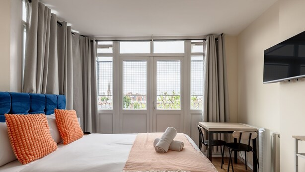 A hotel room with a large bed, a desk, a chair, a TV, and a view of the city through the windows. - Warm Apartments in Queensway (London)