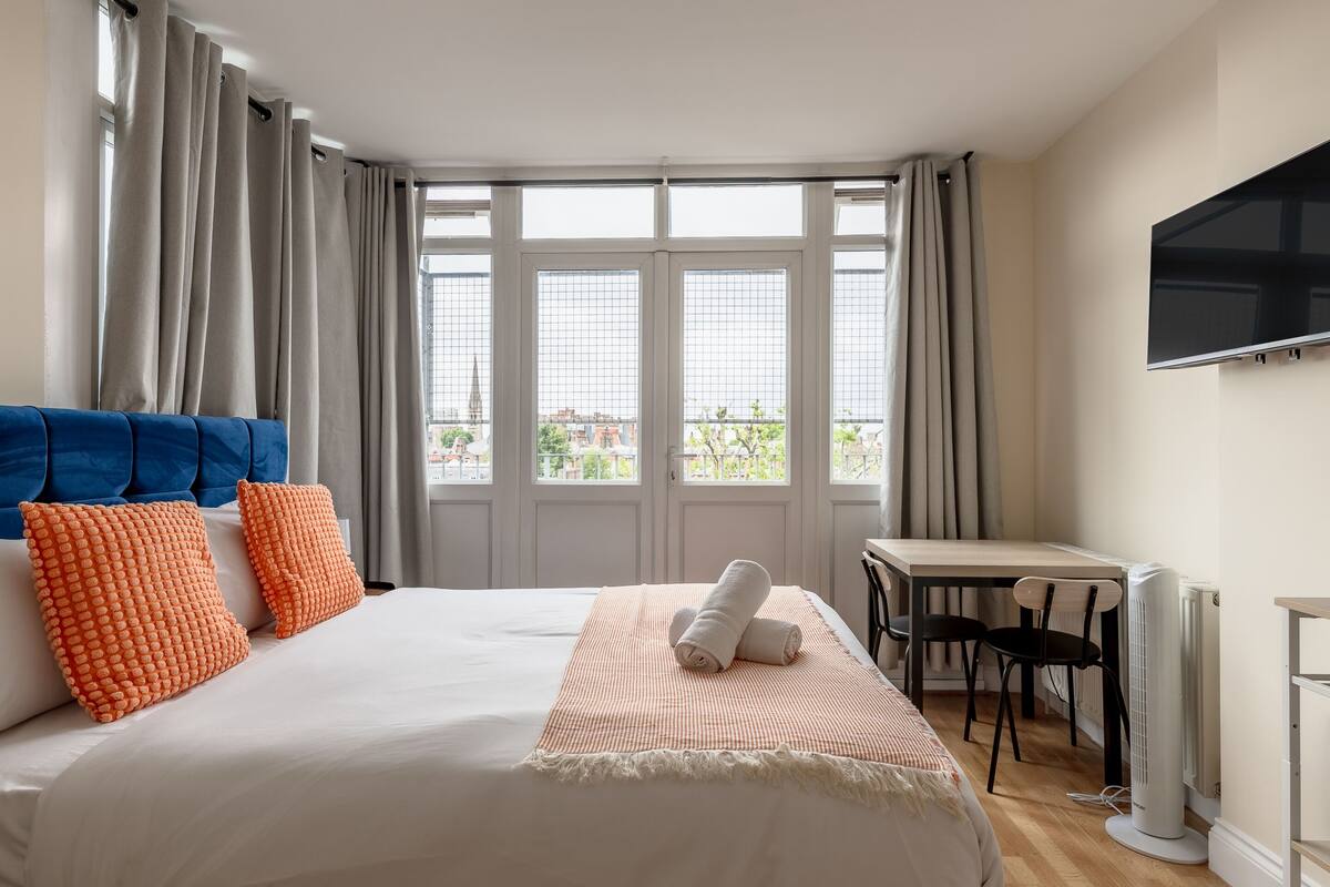 A hotel room with a large bed, a desk, a chair, a TV, and a view of the city through the windows.