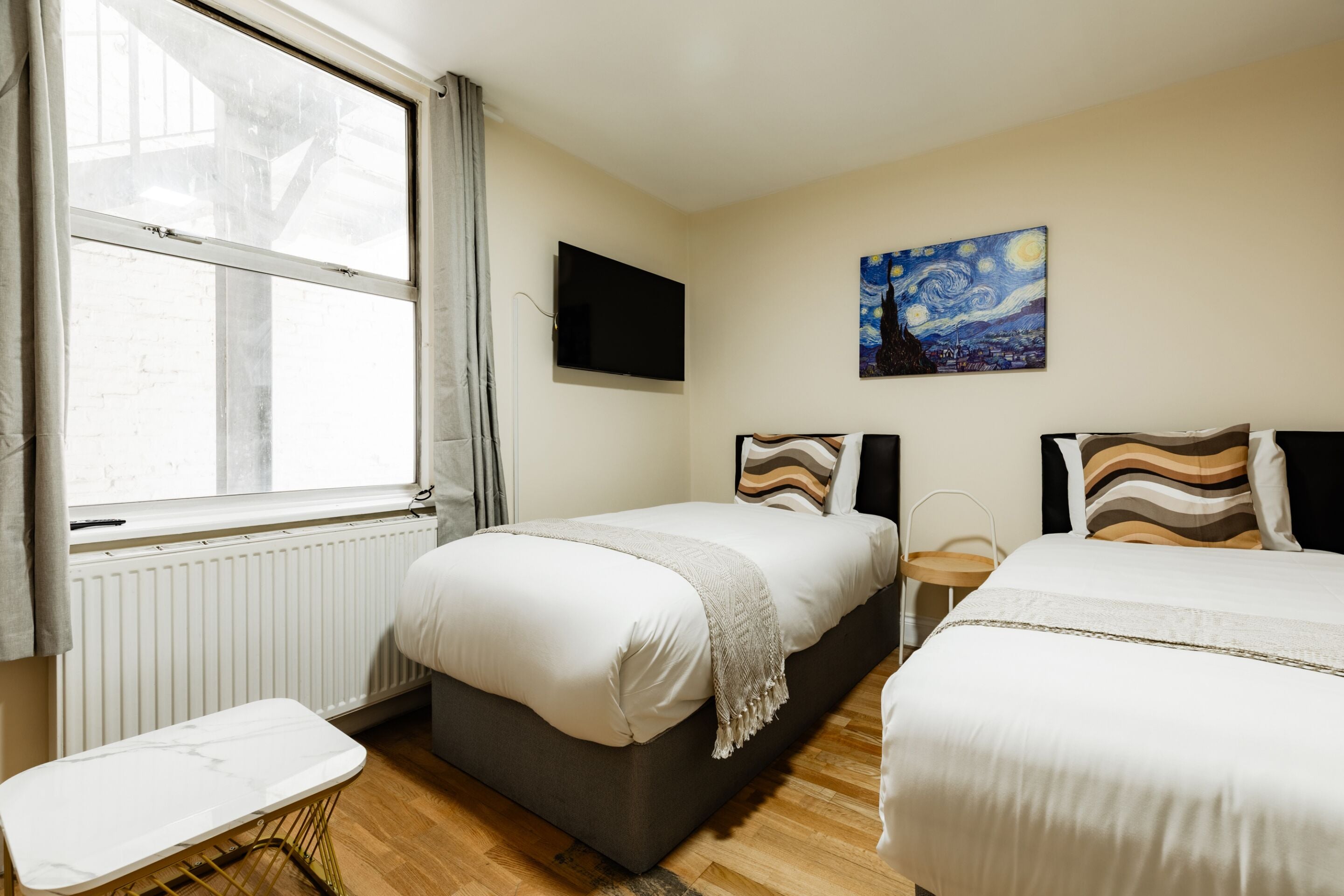 A hotel room with two beds, a flat-screen TV, a painting on the wall, and a window with curtains.