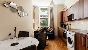 A compact kitchen with a dining area, a washing machine, and a large window with curtains. - Warm Apartments in Queensway (London)
