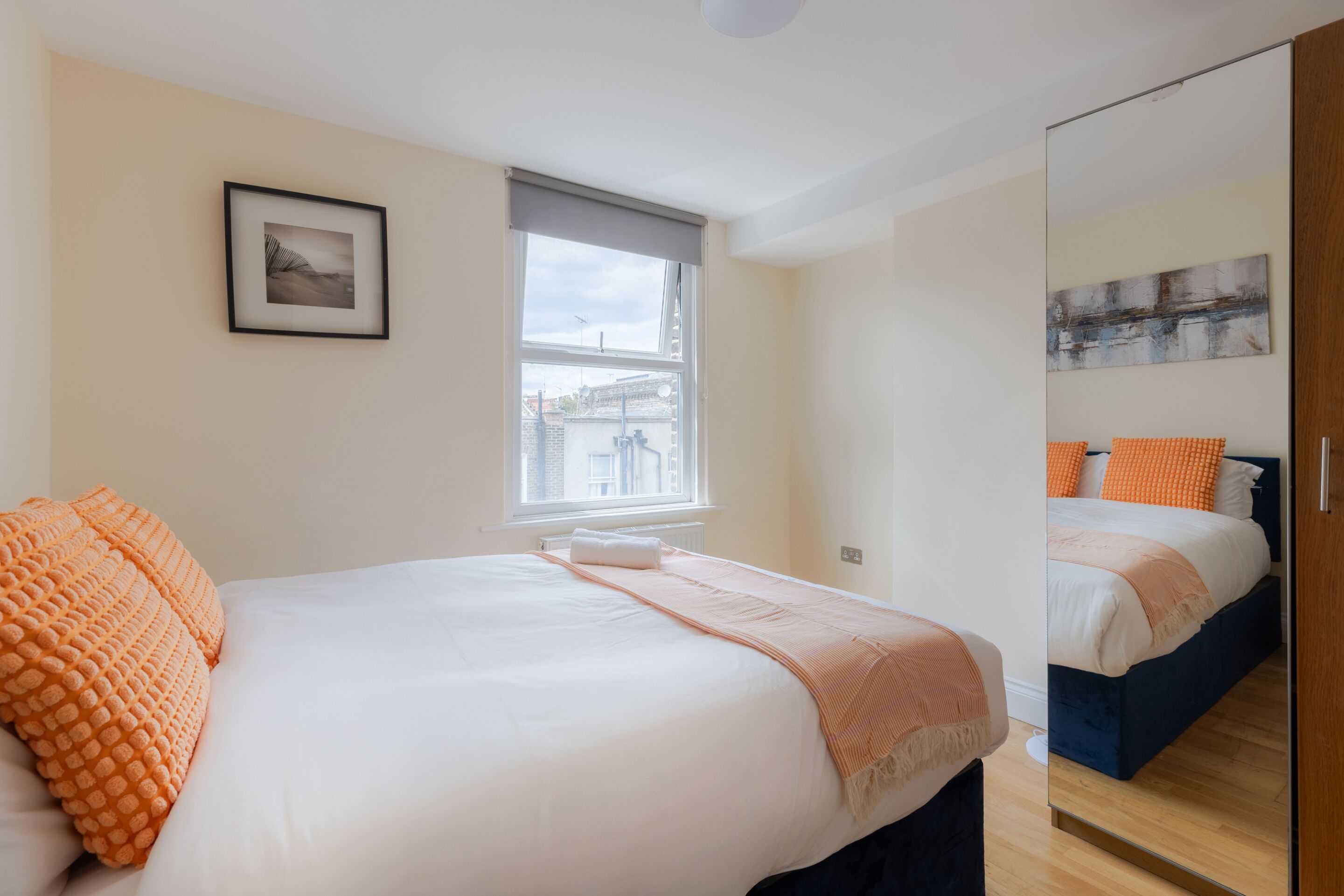 A bedroom with a large bed, a window, a framed picture on the wall, and a mirror reflecting the bed.