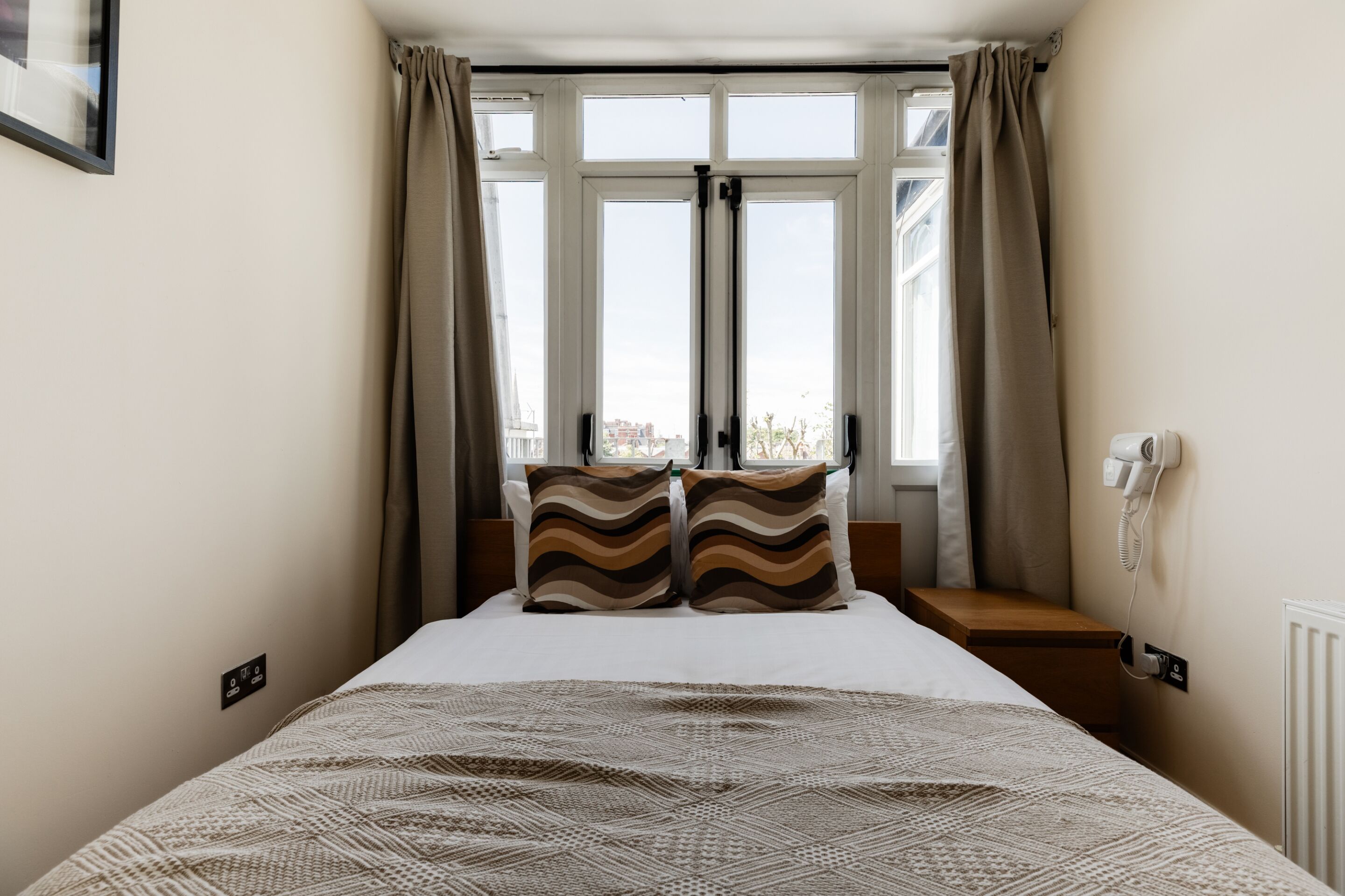 Hotellihuone, jossa on sänky, kaksi tyynyä, ikkuna verhoilla, yöpöytä ja puhelin seinässä.
