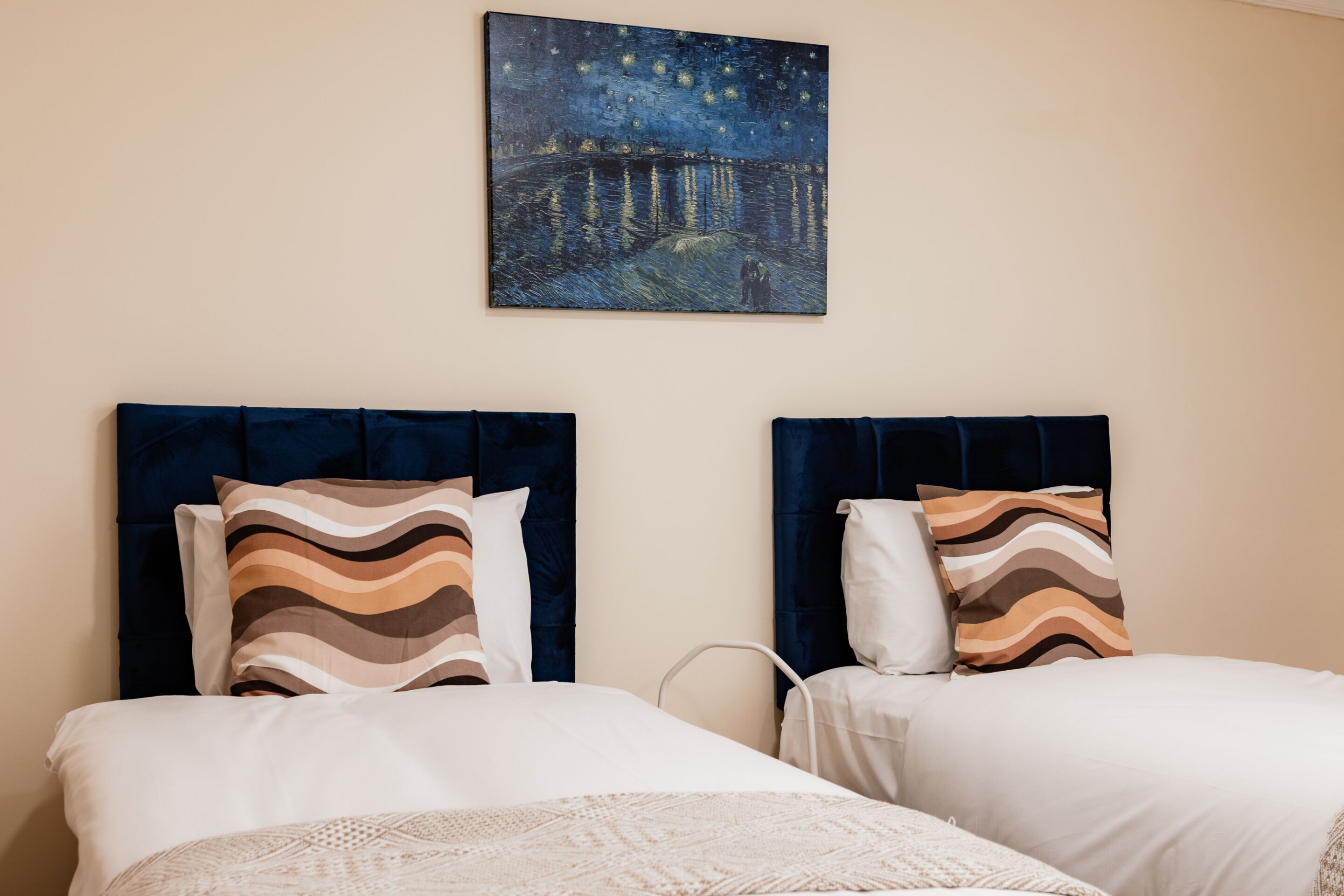 A hotel room with two beds, each with a dark headboard and patterned pillows. A painting of a night scene hangs above the beds.