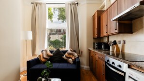 A compact living space with a sofa, a coffee table with a cactus, a window with curtains, a kitchen with wooden cabinets and a Bosch oven. - Warm Apartments in Queensway (London)
