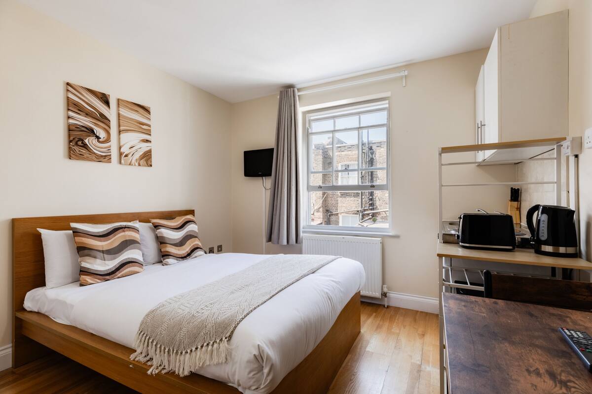 A bedroom with a wooden bed, a TV, a window with a view of buildings, and a kitchenette with a microwave.