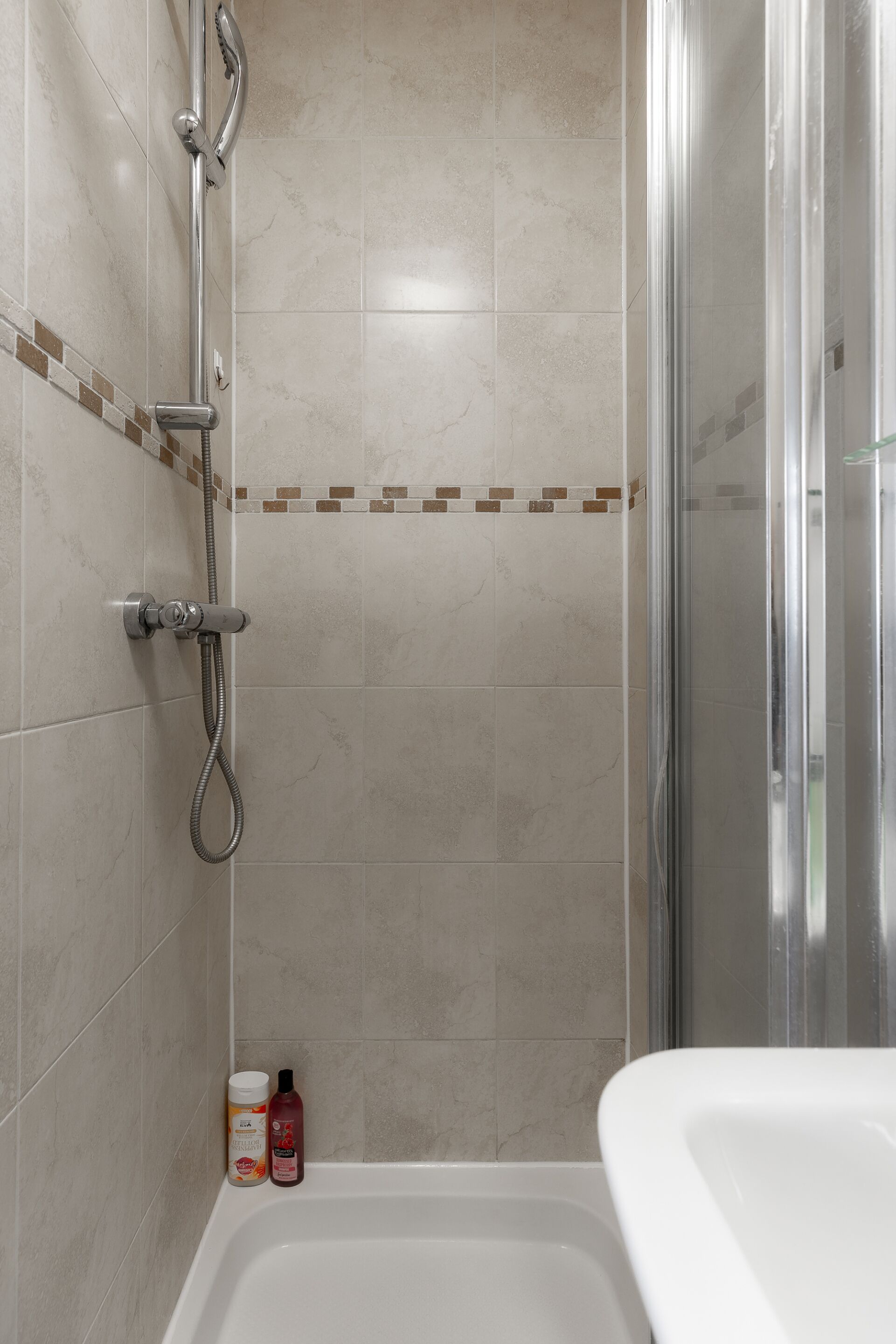A shower area with a glass door, a handheld showerhead, and a wall-mounted faucet. There are two bottles of shampoo and conditioner on the floor.
