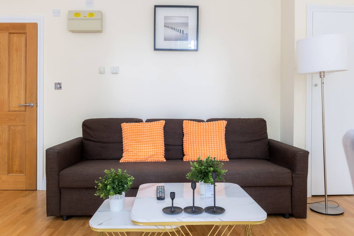 A brown sofa with orange cushions, a white coffee table with a remote, and two potted plants.