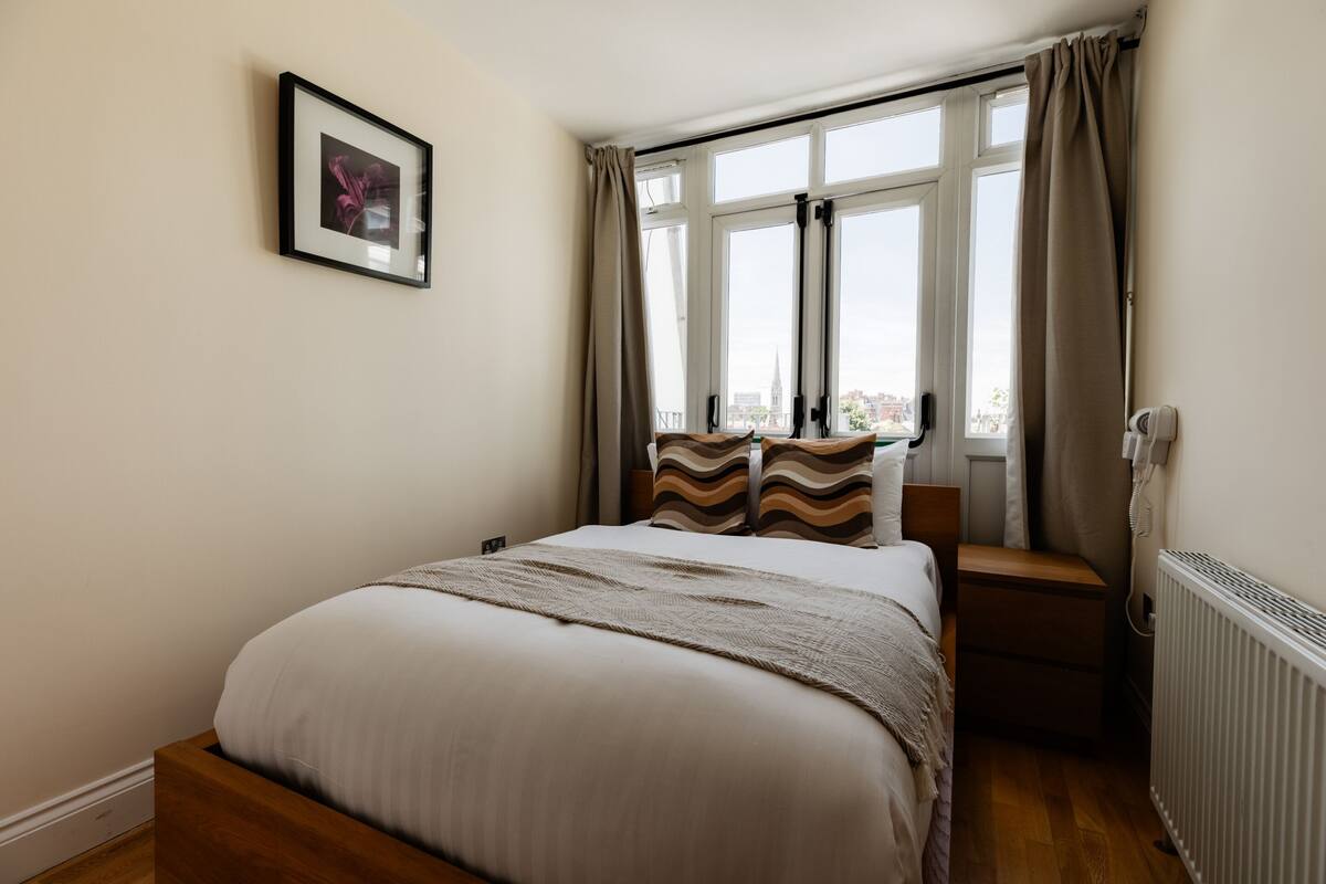 A bedroom with a bed, a window with curtains, a radiator, and a framed picture on the wall.