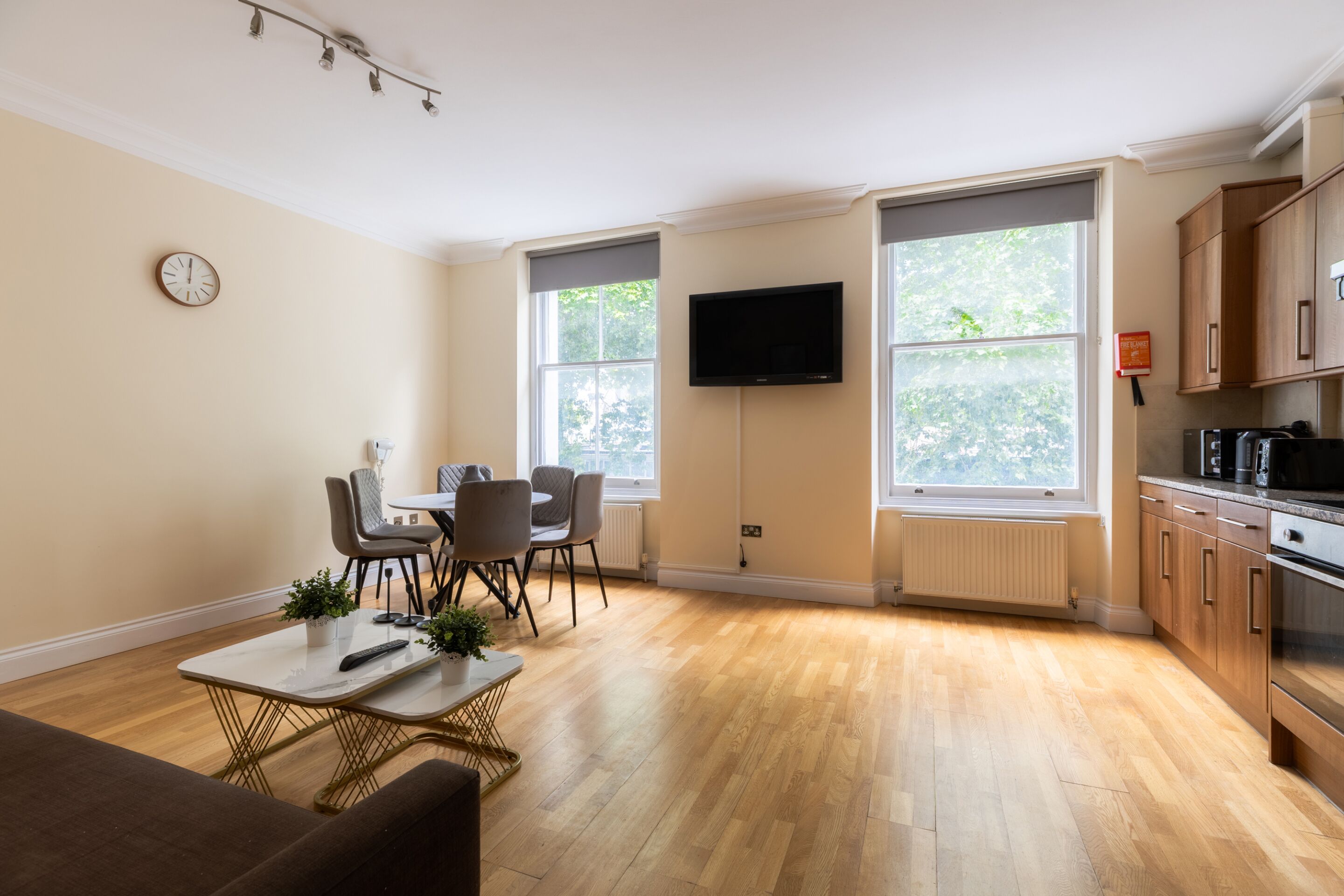 A modern living room with a dining area, wooden flooring, a flat-screen TV mounted on the wall, and a kitchen with wooden cabinets and appliances.