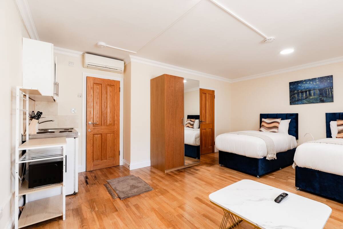 A hotel room with a wooden floor, a white desk with a computer, a bed with a blue headboard, and a painting on the wall.