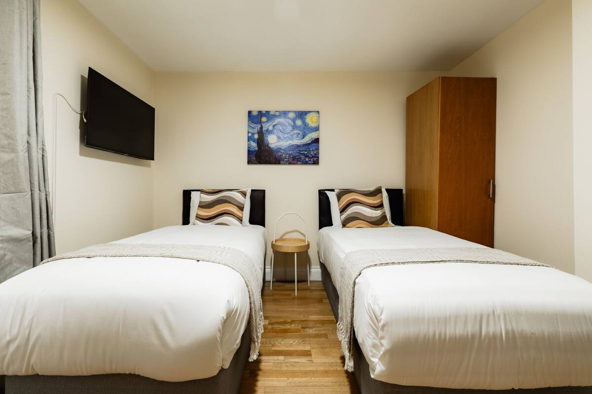 A hotel room with two beds, a flat-screen TV mounted on the wall, a wooden wardrobe, and a painting of a night sky.