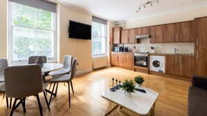 A modern kitchen with wooden cabinets, a built-in oven, and a washing machine. There is a dining area with a table and chairs, and a TV mounted on the wall. - Warm Apartments in Queensway (London)