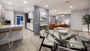 A modern kitchen with white cabinets, a dining area with a glass table, and a living room with a sofa and a coffee table.