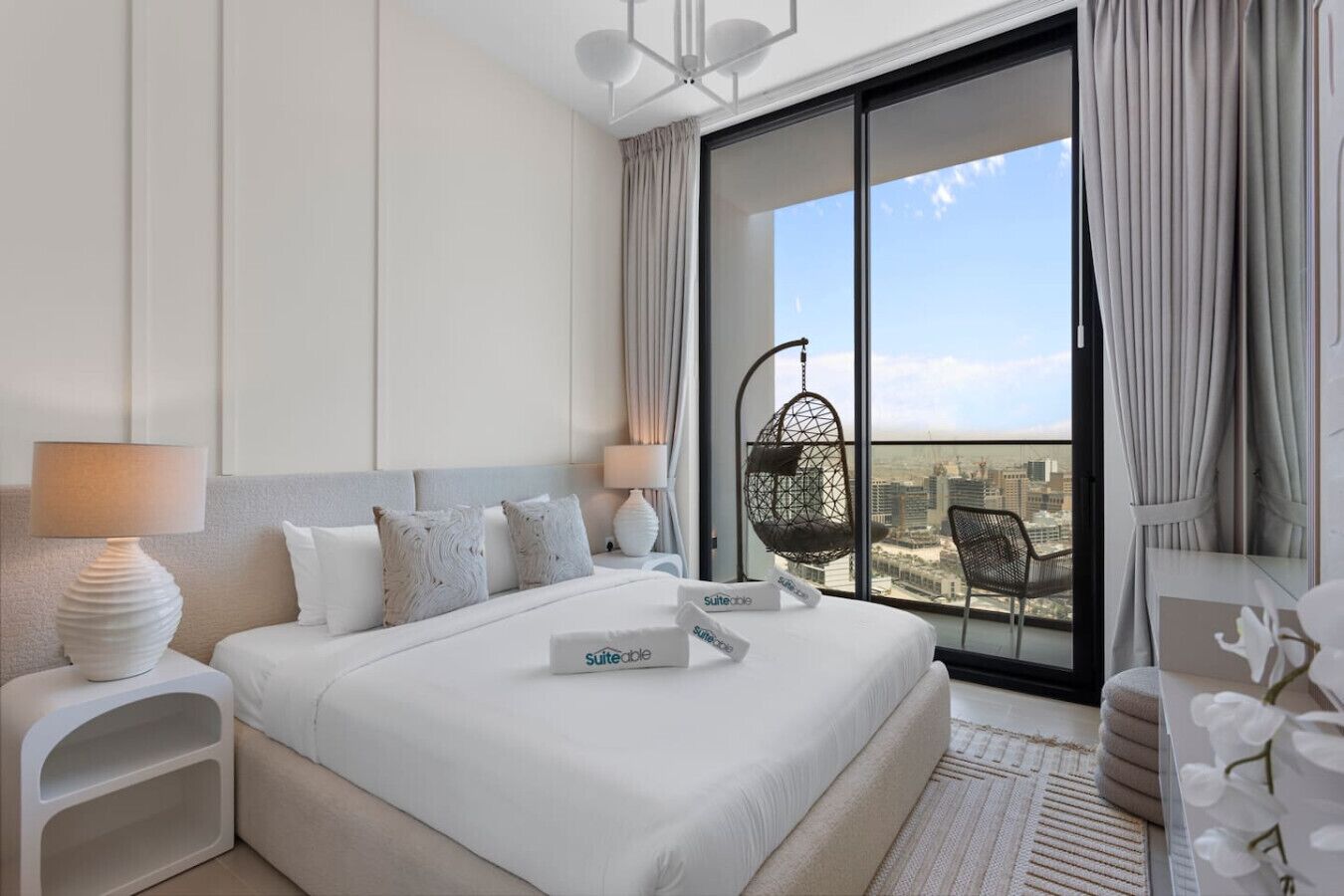 A modern hotel room with a large bed, a hanging chair, and a balcony with a city view.