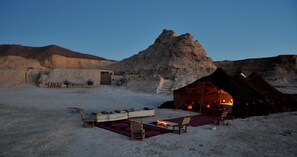 A tented camp with a fire, a thatched-roof structure, and a rocky landscape.