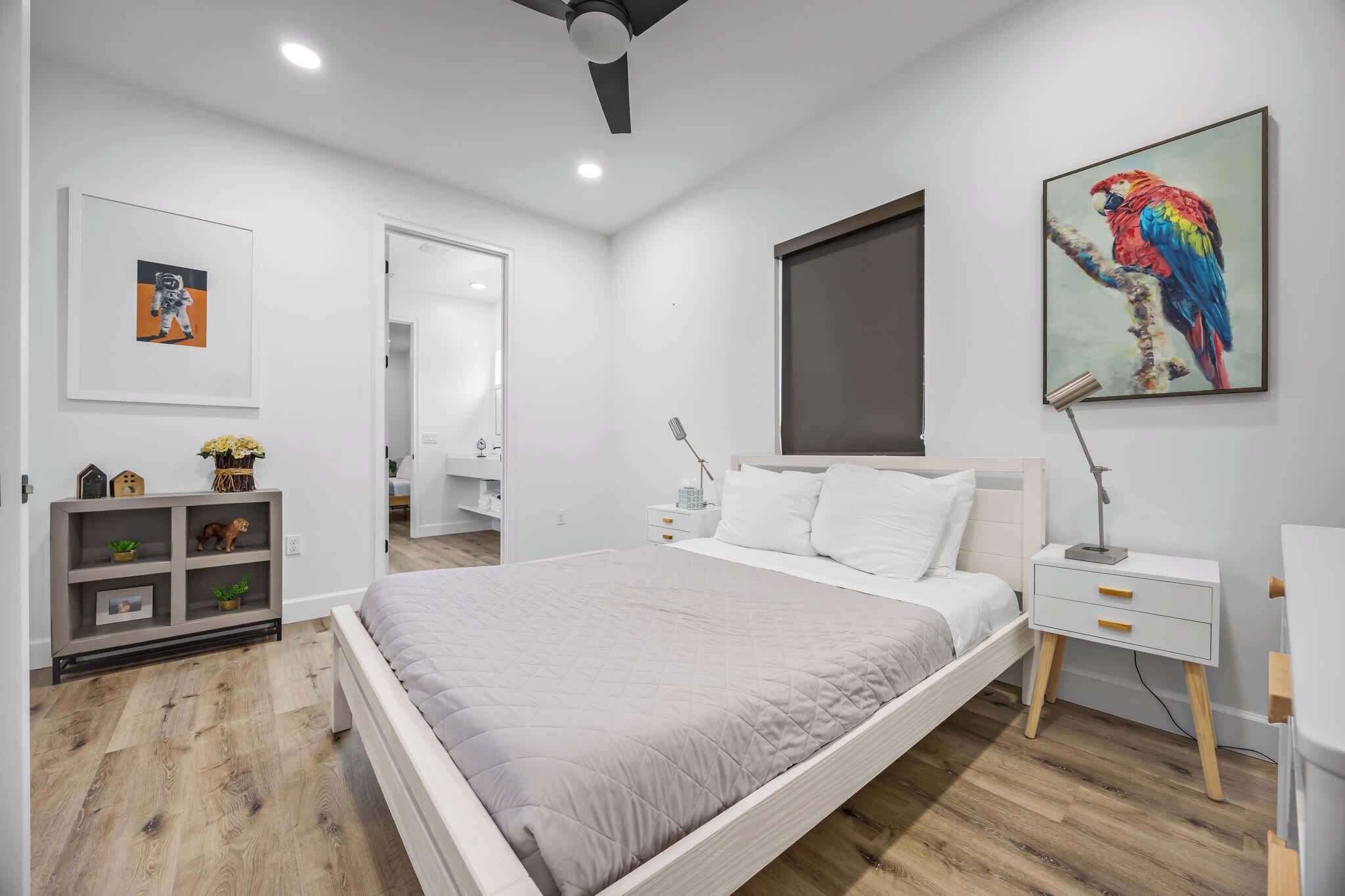 A modern bedroom with a large bed, bedside tables, a nightstand, a ceiling fan, a wall-mounted TV, and a framed artwork of a parrot.