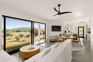 Un salon moderne doté d’une grande porte-fenêtre coulissante donnant sur un paysage désertique, d’un ventilateur de plafond, d’un téléviseur à écran plat et d’un coin repas avec une table ronde et des chaises.