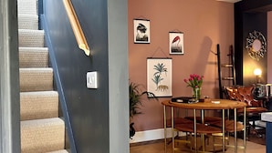 Un escalier dont les marches sont recouvertes de moquette mène à une pièce dont un des murs est brun, ornée d’œuvres d’art encadrées, et qui contient une table avec des chaises et un canapé en cuir.