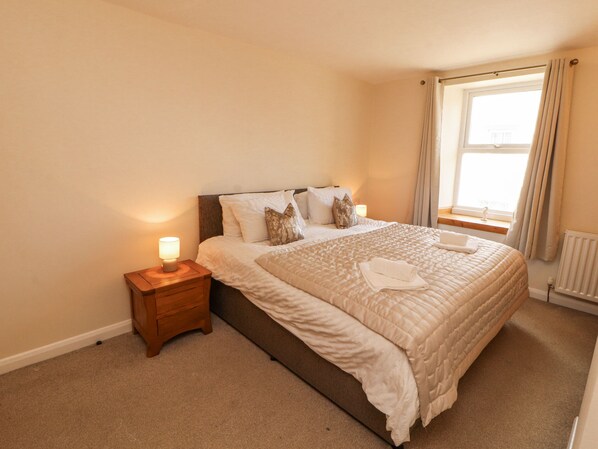 A neatly made bed with a quilted blanket, a bedside lamp, and a window with curtains. - Sun House (Maryport)