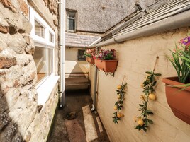 Un estrecho callejón con un muro de piedra, una puerta de madera y una ventana. Hay macetas de terracota con flores y una planta con frutos amarillos en la pared.