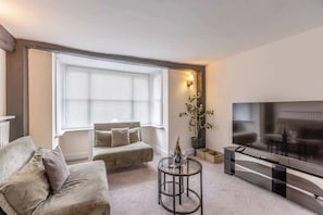 A living room with a sofa, a coffee table, a TV, and a window with blinds. - Charming 3BR Cottage Near Windsor - By Tempstay (Datchet)