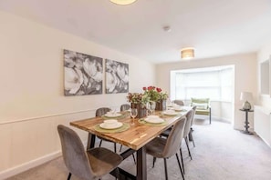 Spiseplads med et træbord dækket til fire personer, omgivet af stole. To billeder hænger på væggene, en vase med røde blomster og en lampe på et sofabord.