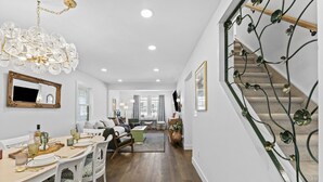 A spacious living area with a dining table set for four, a staircase with decorative metal railings, and a large chandelier.