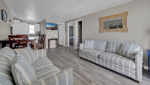 Una sala de estar con un sofá, una mesa de comedor con sillas, una zona de cocina y un pasillo que conduce a otra habitación.