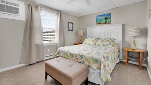 Un dormitorio con cama, banqueta, mesita de noche, ventilador de techo y ventana con cortinas.
