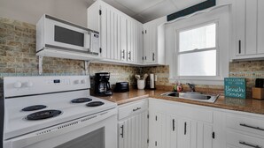 Una cocina con armarios blancos, un microondas, una estufa y una cafetera. Hay una ventana y un letrero que dice 'Welcome to our Beach House'.