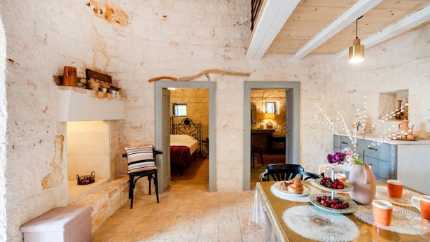 Un interior rústico con paredes de piedra vista, techo de madera, un rincón acogedor para comer con una mesa dispuesta para dos, y una cama en la habitación contigua.