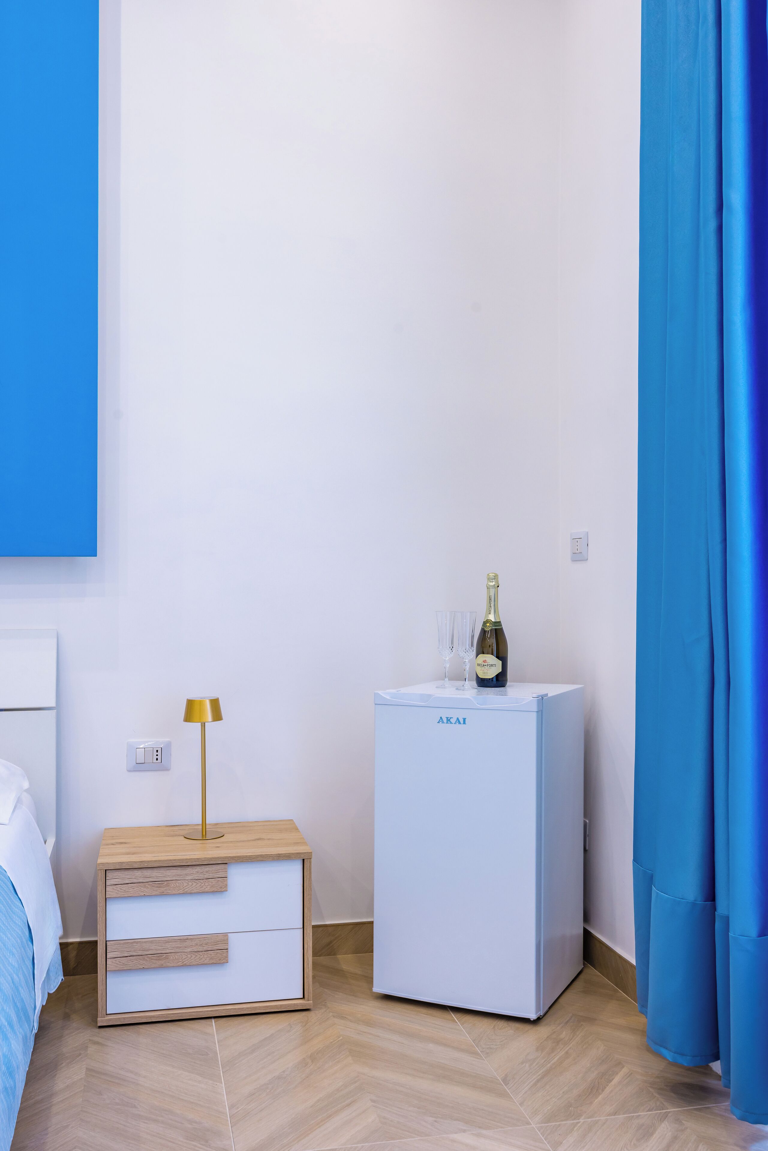 Une chambre avec un lit, une table de chevet avec une lampe, un mini-réfrigérateur et un rideau.