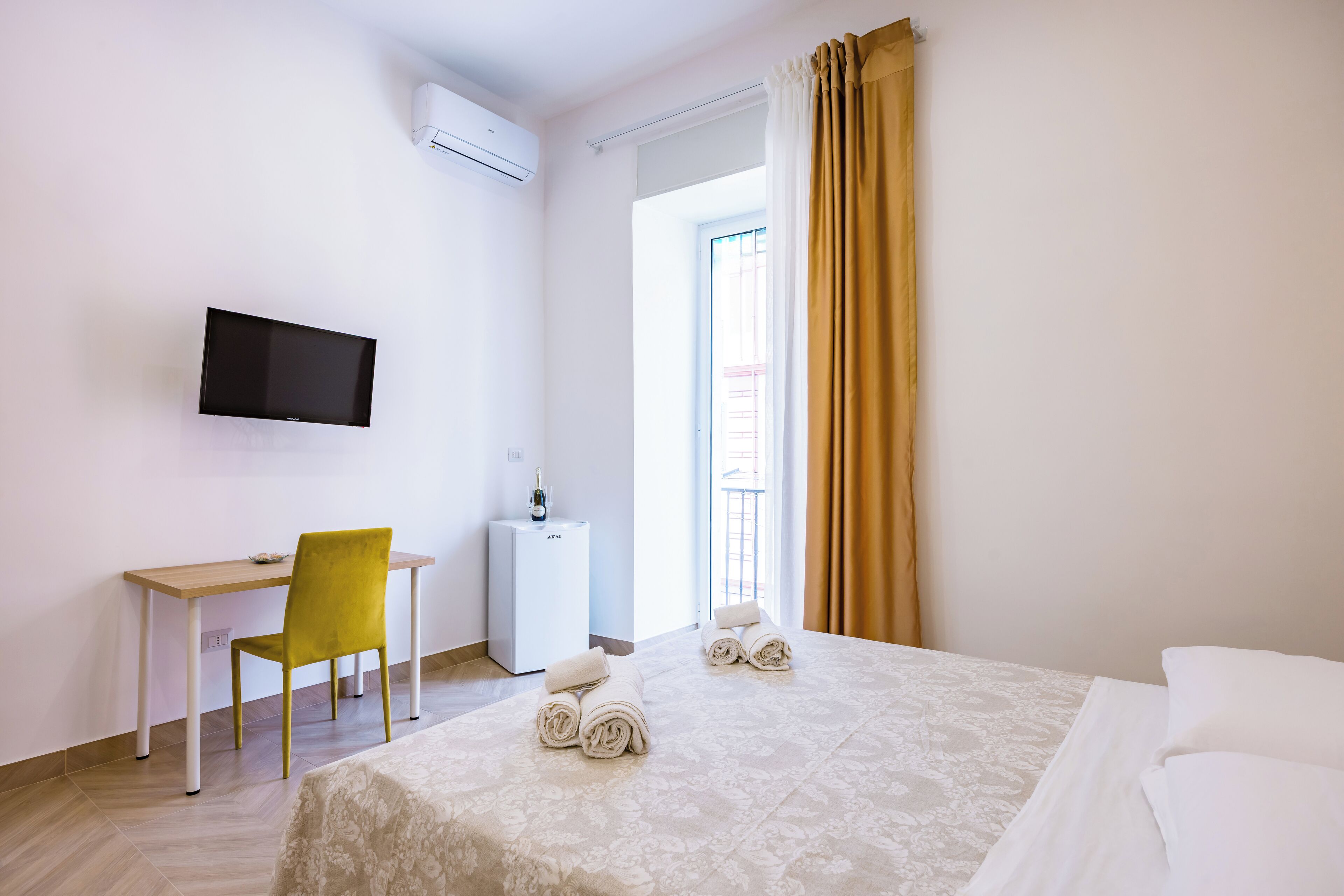 A hotel room with a bed, a desk, a chair, a TV, a minibar, and a window with curtains.