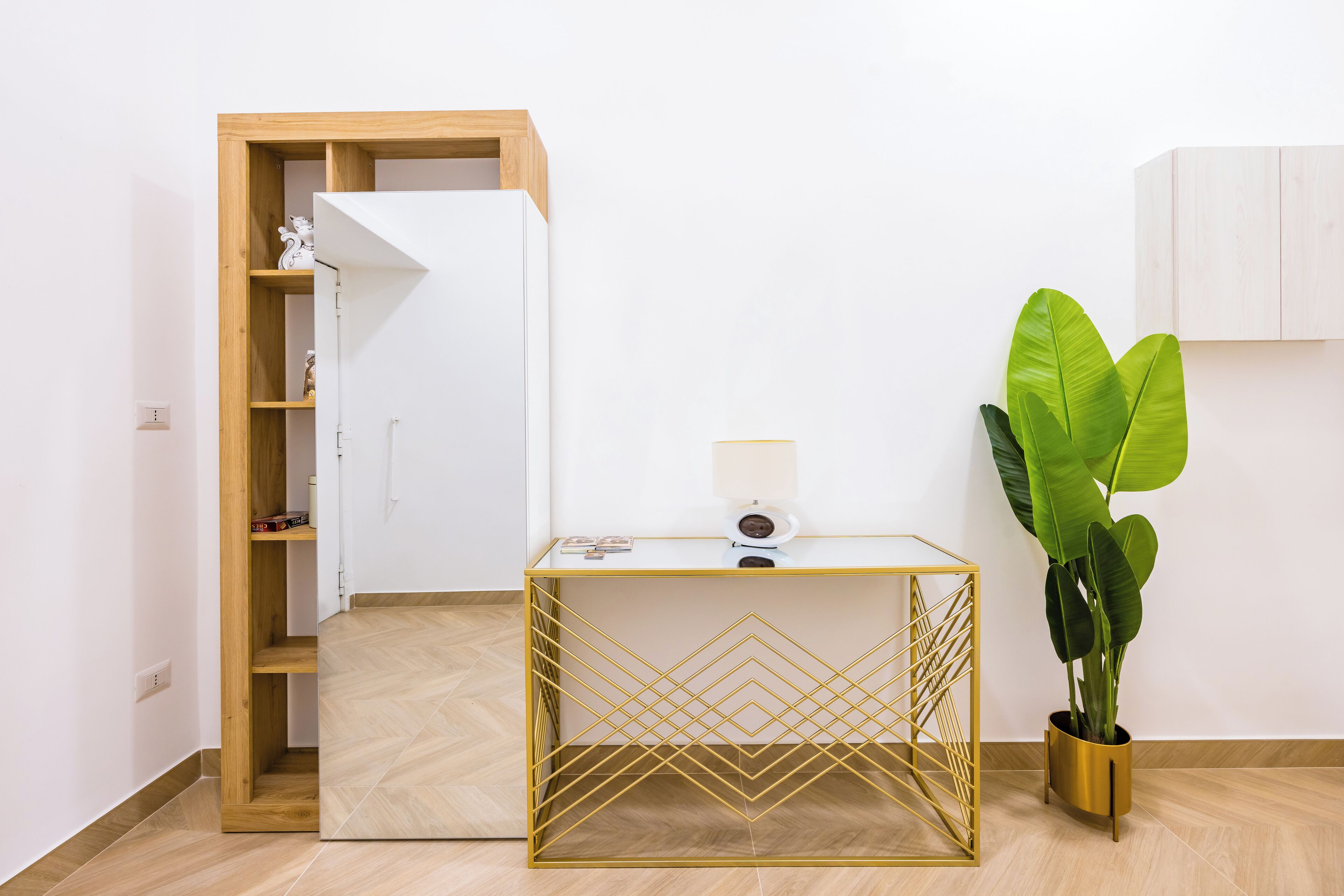 A room with a wooden wardrobe, a glass table with a geometric gold frame, a potted plant, and a white cabinet.
