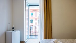 A hotel room with a bed, a small white minibar, and a view of a building through an open door. - Essentia Home (Naples)