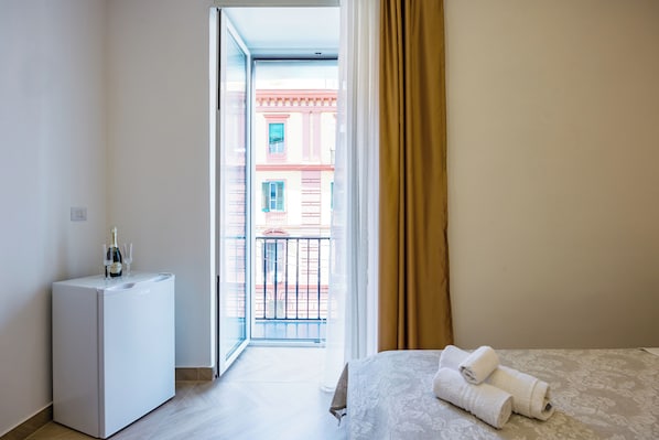Une chambre d’hôtel avec un lit, un petit minibar blanc et une vue sur un immeuble à travers une porte ouverte.