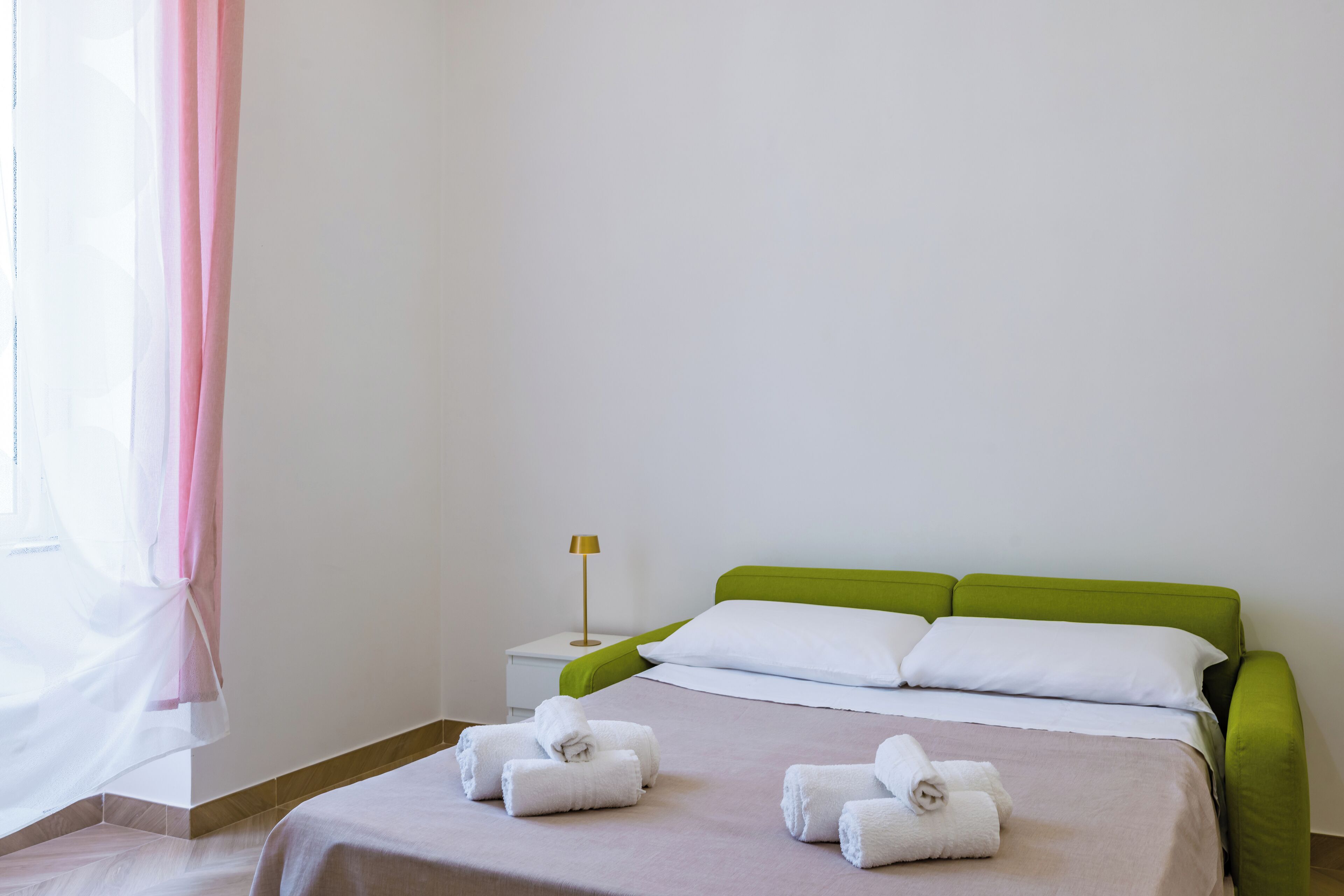 Una cama bien tendida con cabecera verde y ropa de cama blanca, una lámpara de noche y toallas enrolladas sobre la cama.