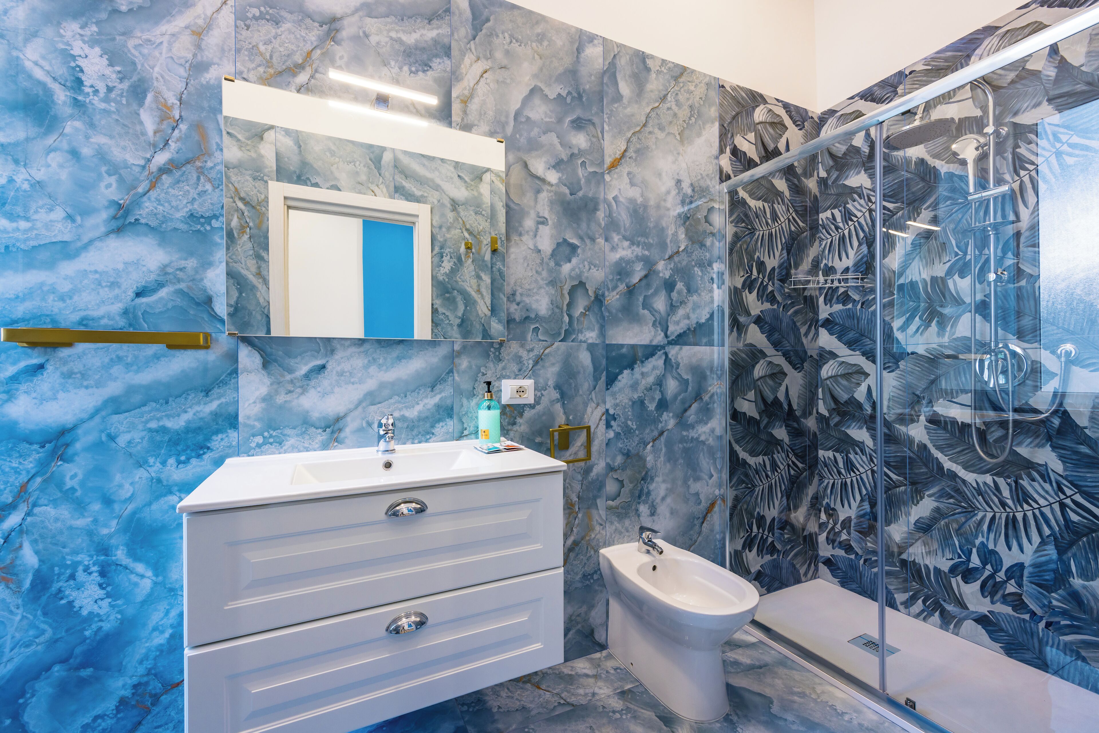 Une salle de bain avec un mur en marbre, un meuble-lavabo blanc avec des tiroirs, un évier, un bidet et une cabine de douche en verre.