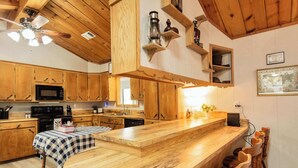 A kitchen with wooden cabinets, a microwave, a stove, a checkered tablecloth, and a bar area with wooden chairs. - Waterfront Lake Retreat • Private Dock & Fire Pit (Anacoco)