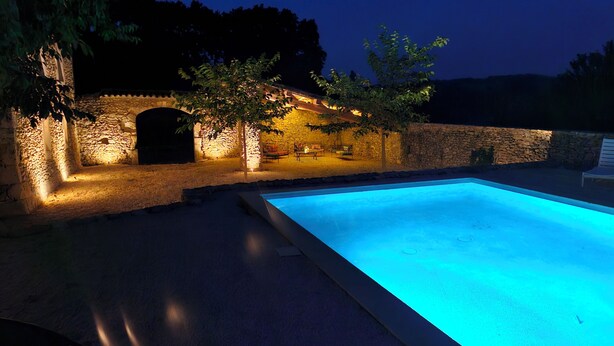 A swimming pool with a stone wall and a small archway, surrounded by trees and illuminated by warm lighting. - Le Mas Tillet (Goudargues)