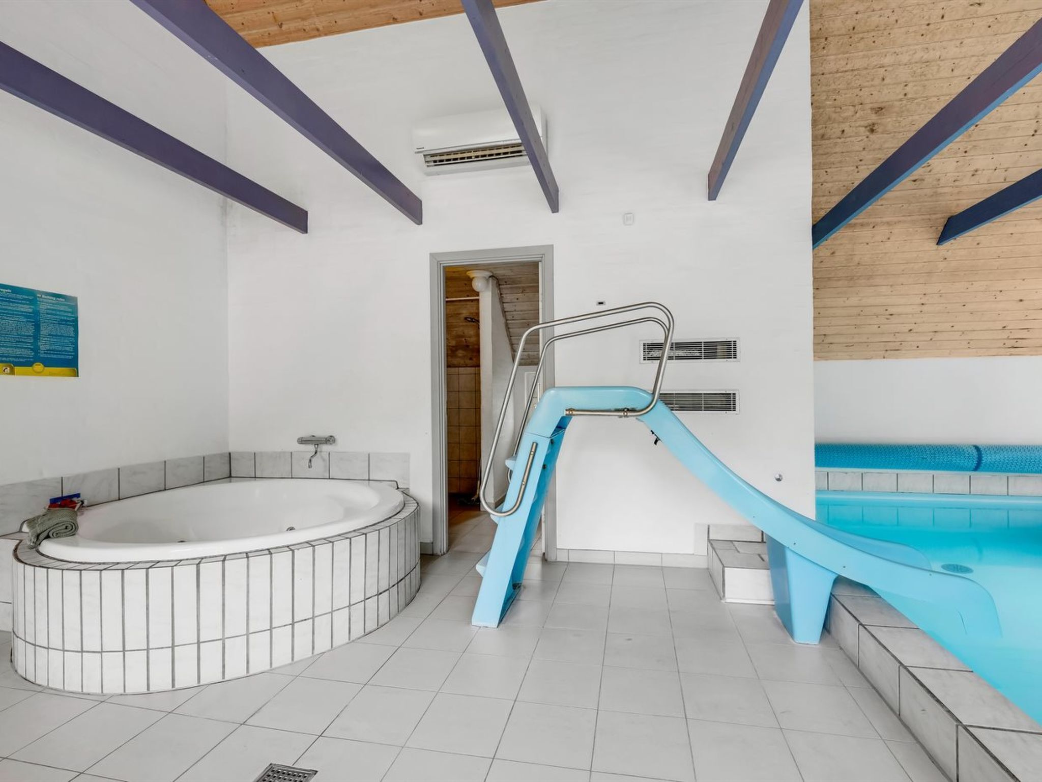 Un espace piscine intérieur moderne, doté d’un jacuzzi, d’un toboggan bleu et d’un escalier menant à une zone de douche.