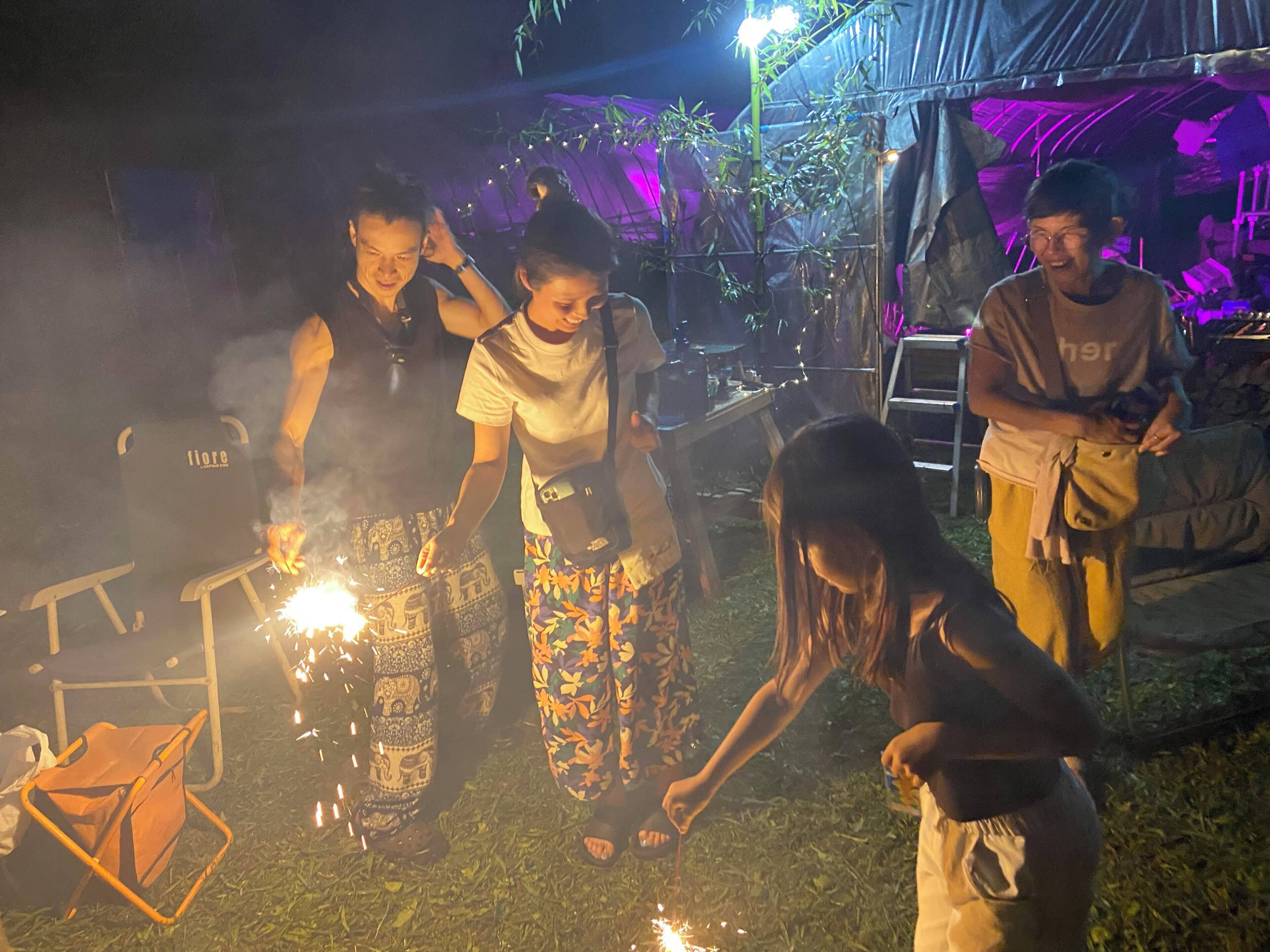A group of people gathered around a campfire with sparklers, in an outdoor setting with tents and foliage in the background.