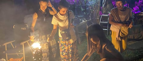 A group of people gathered around a campfire with sparklers, in an outdoor setting with tents and foliage in the background.