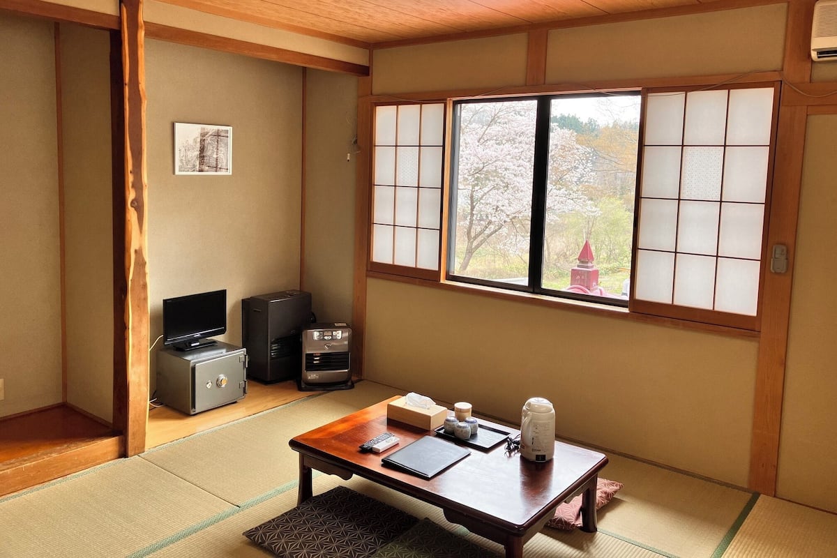 A traditional Japanese room with tatami flooring, a sliding door, a television, a speaker, and a view of cherry blossoms through a shoji screen.