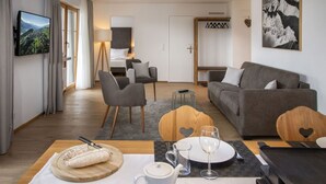 A modern living room with a sofa, chairs, a dining table set for two, and a TV mounted on the wall.