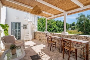 A covered outdoor seating area with stone walls, a white building with a blue door, and a view of greenery.