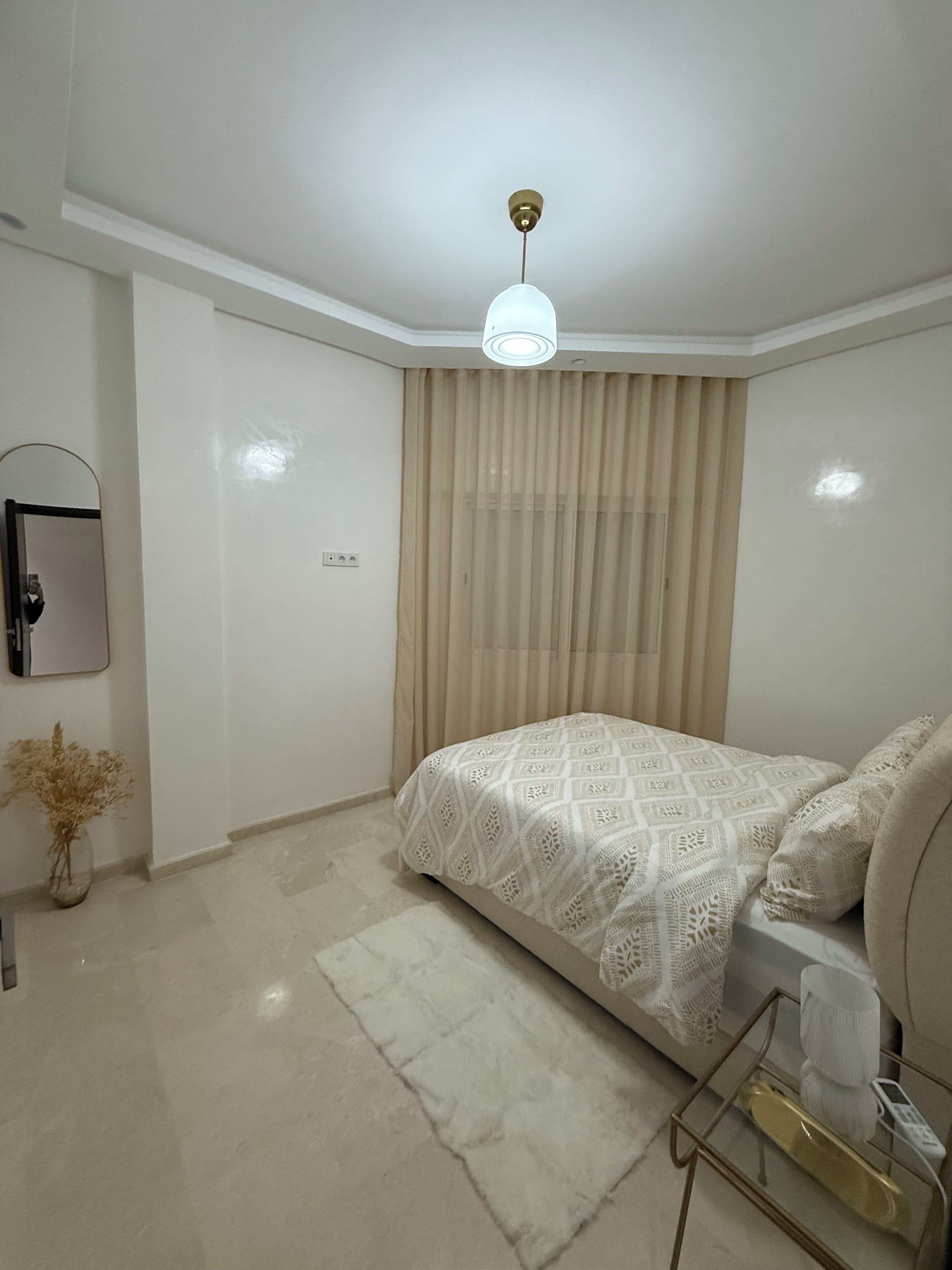 A bedroom with a bed, a bedside table, a lamp, a mirror, and a window with curtains.